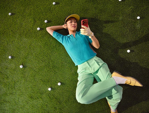 Una persona tumbada boca arriba en un césped salpicado de pelotas de golf, mirando un smartphone.