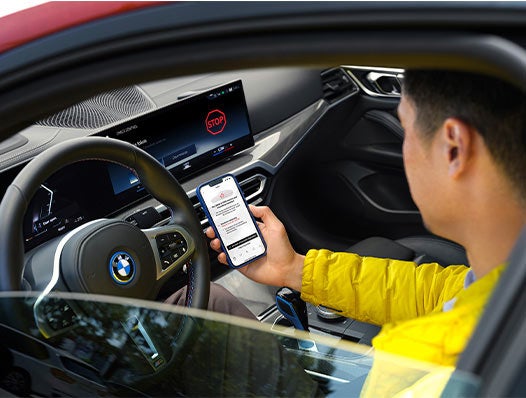 Un hombre sostiene un teléfono inteligente con la pantalla visible mientras está sentado en el asiento del conductor de un BMW.